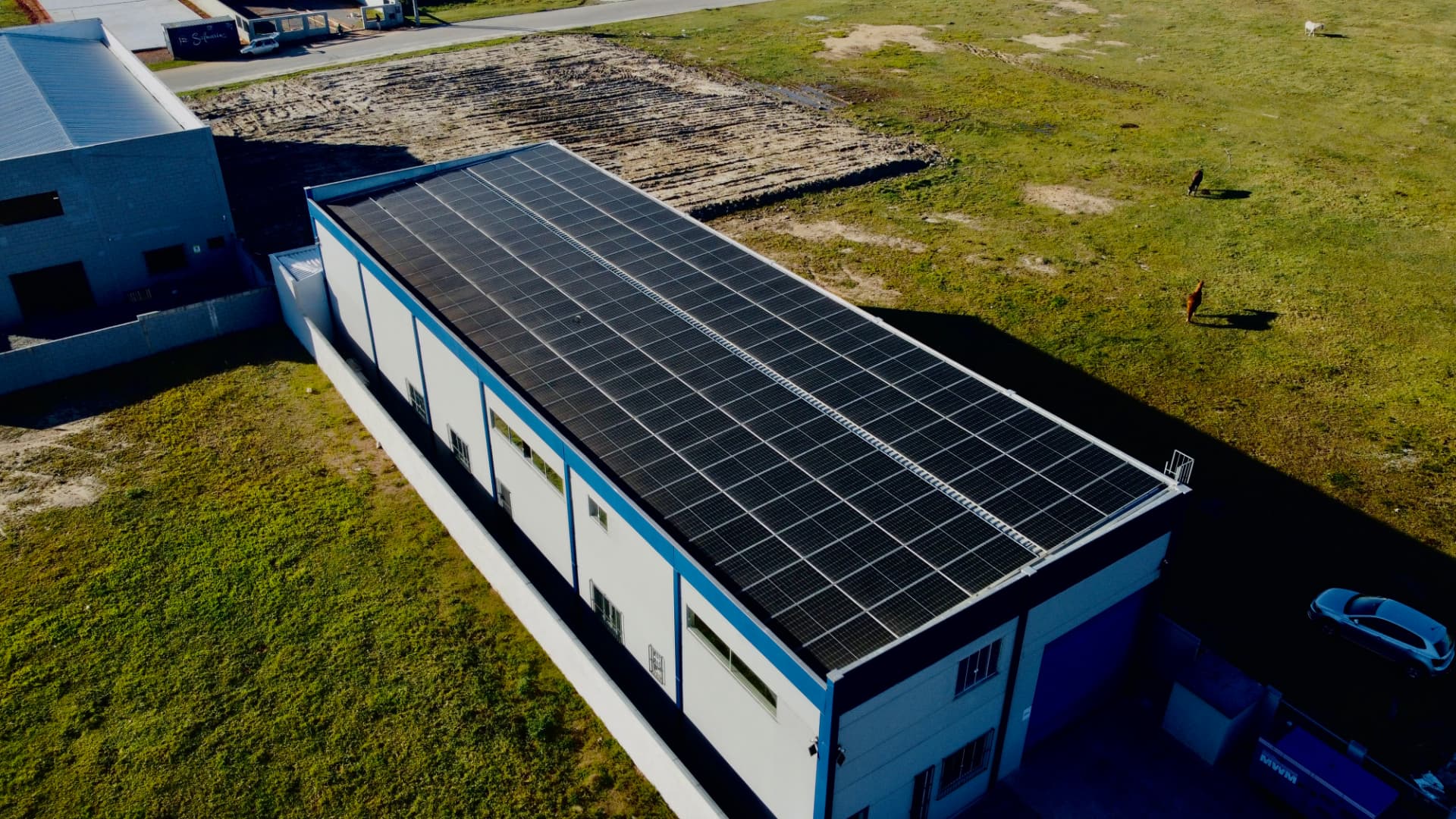 Vista aérea de galpão comercial com telhado coberto por módulos fotovoltaicos, em terreno amplo com campo gramado
