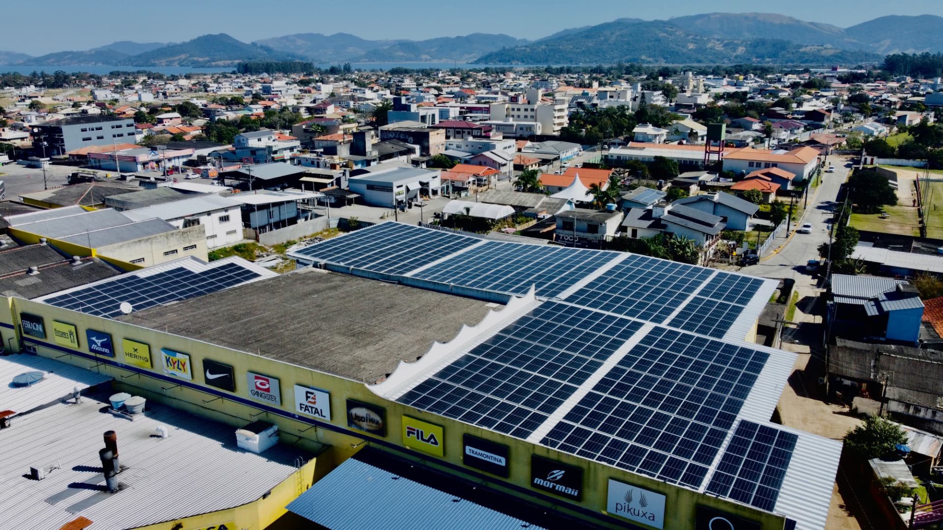 Vista aérea de telhado comercial coberto por módulos fotovoltaicos, com a cidade ao fundo